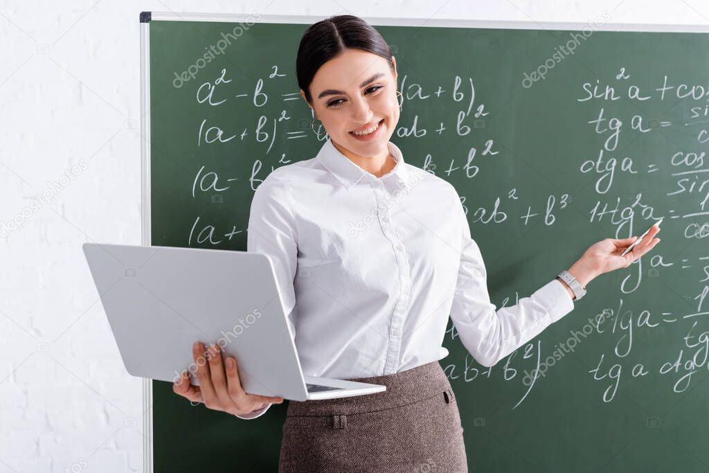 Smiling teacher having video call on laptop near chalkboard in classroom 