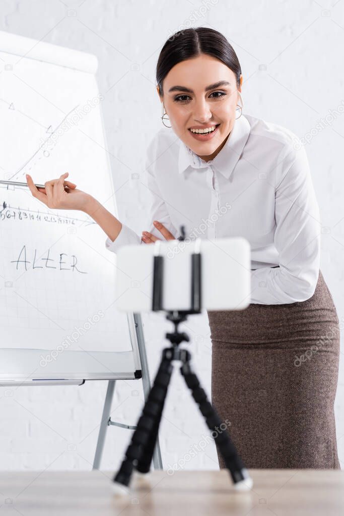 Smiling businesswoman in earphones pointing at flipchart near blurred smartphone during video call 