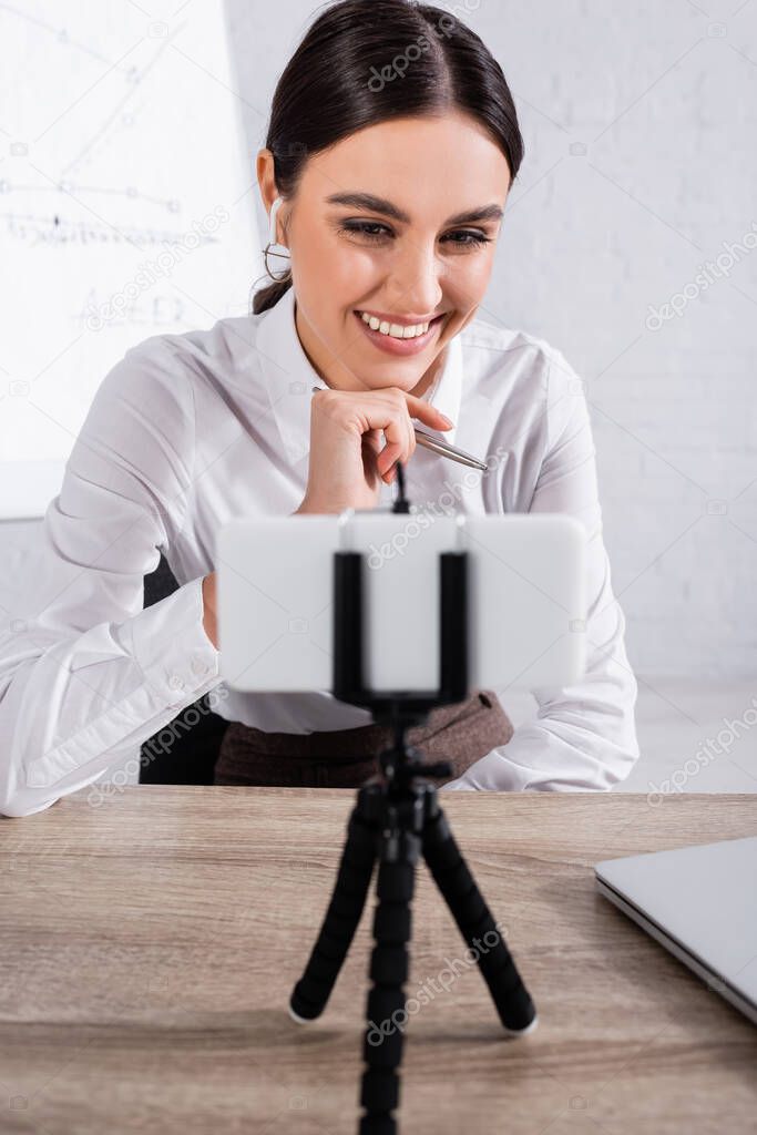 Cheerful businesswoman in earphone having video chat on blurred smartphone in office 