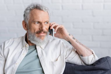 Dövmeli ve sakallı bir adam, gri saçlı, akıllı telefondan konuşuyor. 