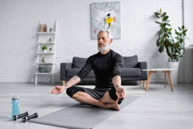 Oturma odasındaki dambılların yanında yoga minderi üzerinde meditasyon yapan gri saçlı bir adam.