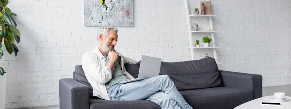 Skäggig Man Tittar Laptop Medan Sitter Soffan Banner — Stockfoto