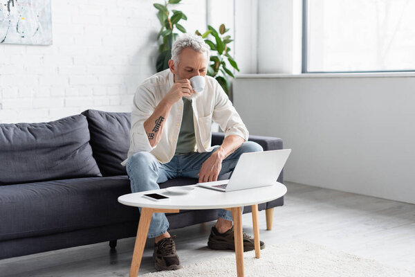 bearded man drinking coffee and using laptop near smartphone with blank screen on coffee table