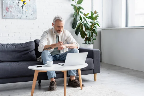 Skäggig Man Pekar Laptop Medan Sitter Soffan Nära Kopp Kaffe — Stockfoto