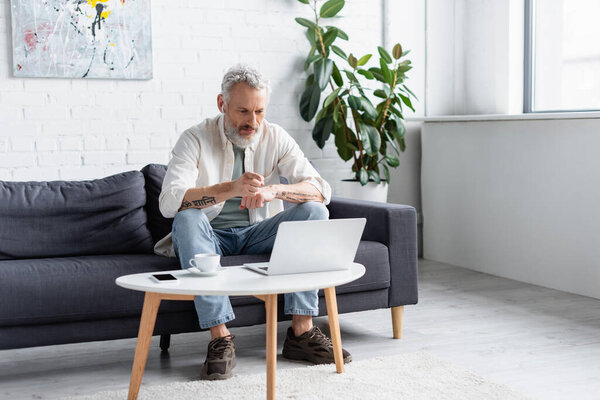 bearded man pointing at laptop while sitting on couch near cup of coffee and smartphone with blank screen on coffee table
