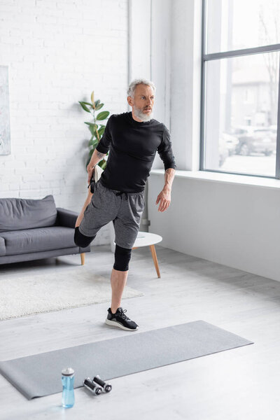 full length of bearded and tattooed man in sportswear exercising on fitness mat near dumbbells in living room 