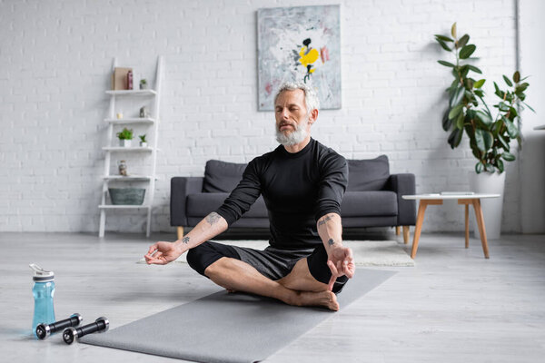 мужчина с седыми волосами медитирует на коврике для йоги рядом с гантелями в гостиной, баннер