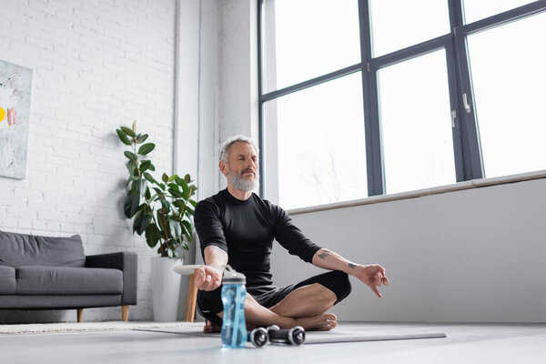 bearded man with grey hair meditating on yoga mat near dumbbells in living room