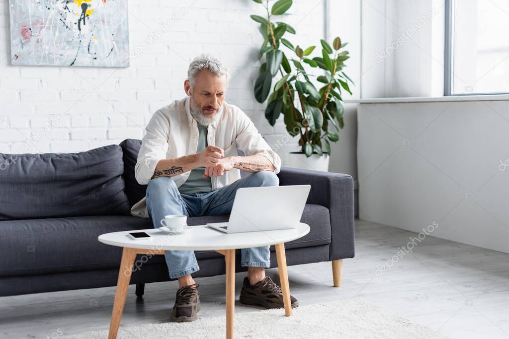 bearded man pointing at laptop while sitting on couch near cup of coffee and smartphone with blank screen on coffee table