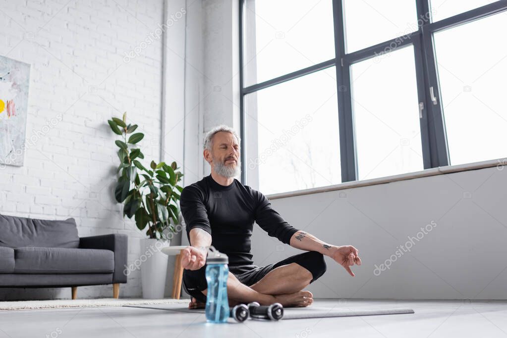 bearded man with grey hair meditating on yoga mat near dumbbells in living room