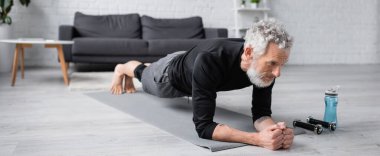 Çıplak ayaklı gri saçlı adam oturma odasındaki dambılların yanında spor tahtası yapıyor.