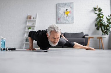 Gri saçlı güçlü adam oturma odasındaki halterlerin yanında spor yapıyor.
