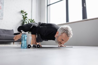 Oturma odasındaki halterlerin yanında spor yapan gri saçlı güçlü bir adam.