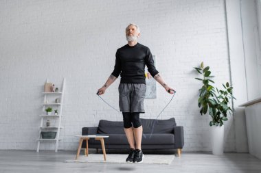 Spor giyimli sakallı adam modern oturma odasında ip atlama egzersizi yapıyor. 