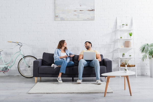 счастливый мусульманский фрилансер, используя ноутбук рядом с подругой с цифровой планшет на диване 