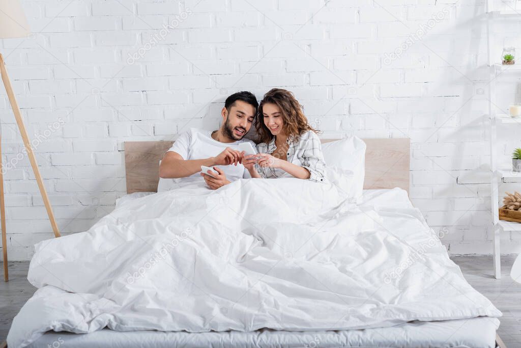 interracial couple smiling while lying on bed and using smartphones 
