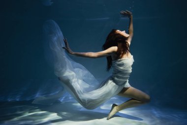 Mavi suyla havuzda yüzen zarif, beyaz elbiseli kadını aydınlatıyorlar.