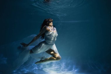 Mavi suyla havuzda yüzen beyaz, zarif elbiseli genç bayanı aydınlatıyorlar. 