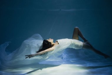 Mavi suyla havuzda yüzen beyaz, zarif elbiseli genç bayanı aydınlatıyorlar. 
