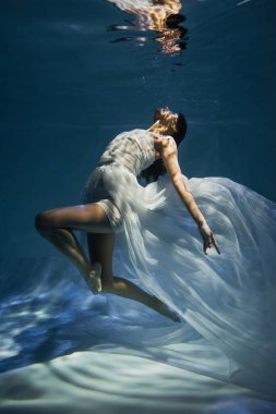 lighting on young graceful woman in white dress swimming in pool with blue water  clipart