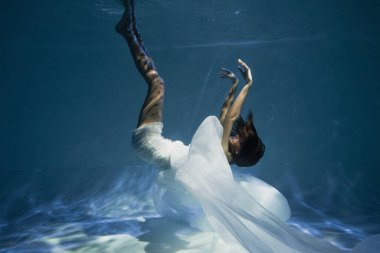 Beyaz elbiseli genç bir bayanı aydınlatmak mavi suyla havuza dalmak. 