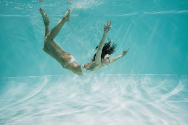 Beyaz mayo giymiş zarif ve esmer bir kadın havuzda yüzüyor. 
