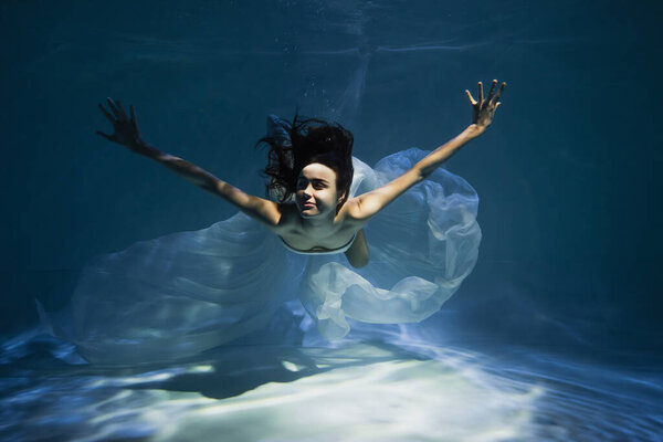 lighting on smiling young woman in white elegant dress swimming in pool 