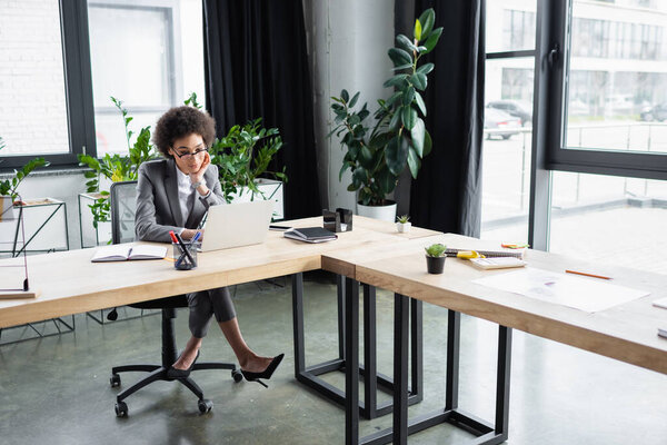 African american manager in eyeglasses looking at laptop near stationery on table 