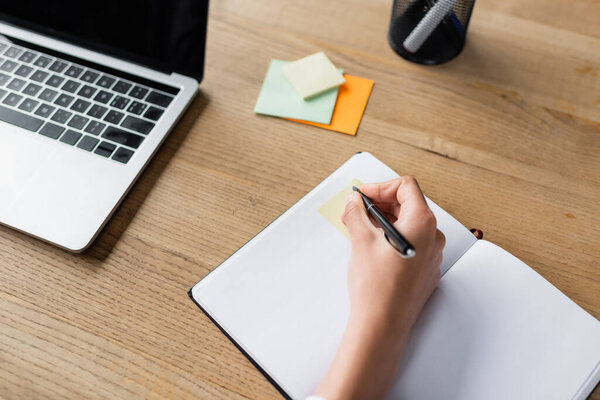 Cropped view of businesswoman writing on copy book near sticky notes and laptop 