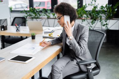 Afrikalı Amerikalı iş kadını cep telefonuyla konuşuyor ve belgenin yanında kalem tutuyor. 