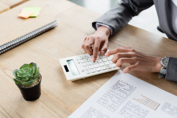 Cropped view of businesswoman using calculator near document and blurred plant 