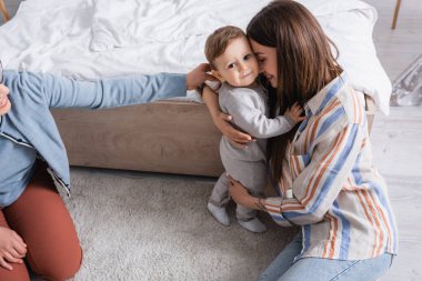 Bebek, yatak odasında gülümseyen annesiyle sarılırken kameraya bakıyor. 