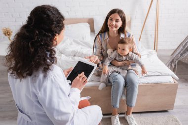 Beyaz önlüklü Afrikalı Amerikalı çocuk doktoru yatak odasında anne ve oğlunun yanında boş ekranlı dijital tablet kullanıyor. 