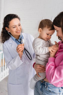 Steteskoplu Afro-Amerikan çocuk doktoru. Mutlu bir annenin ellerinde küçük bir çocuğun yanında.