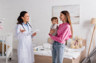 Mutlu anne kollarını tutuyor, bebek çocuğu dijital tabletli Afrikalı Amerikalı çocuk doktorunun yanında. 