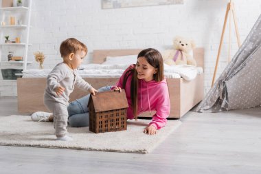 Mutlu anne halının üzerinde bebek oğlunun yanında ev modeline bakıyor. 