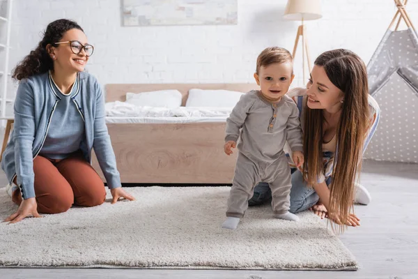 Pleased Interracial Women Looking Infant Boy Standing Carpet — Stock Photo, Image