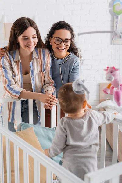 Onnellinen Rotujenvälinen Naiset Etsivät Hämärtynyt Pikkulapsi Poika Vauvan Pinnasänky Makuuhuoneessa — kuvapankkivalokuva