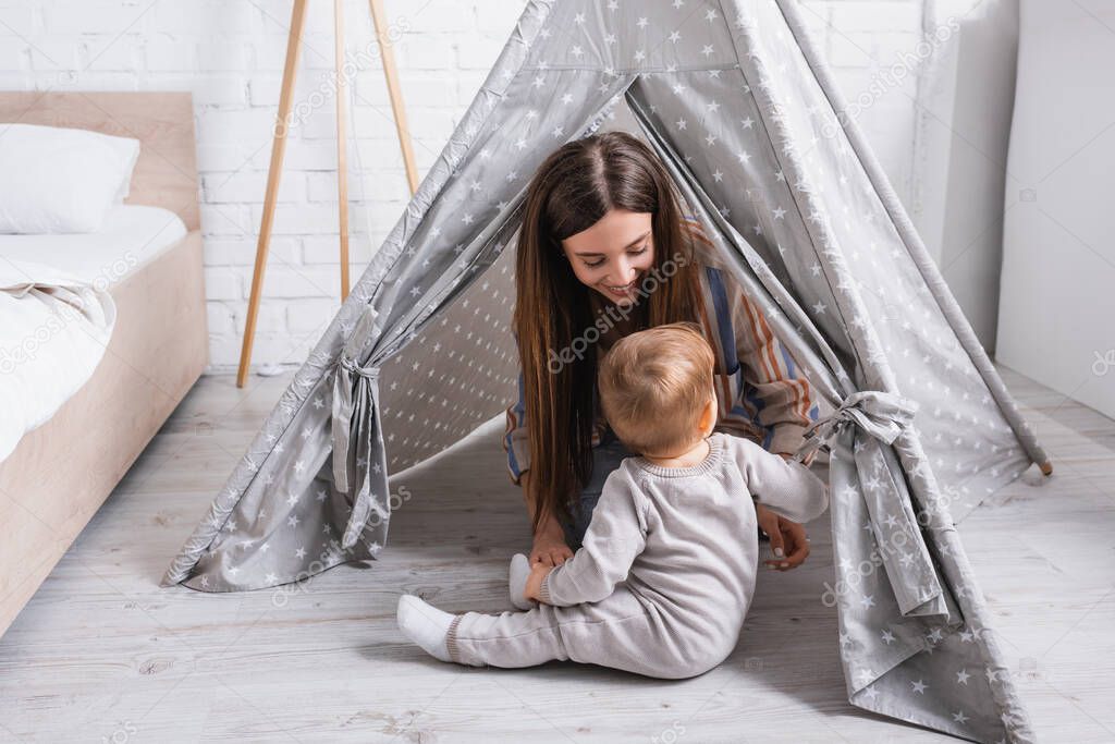 happy mother looking at baby son sitting near tipi in bedroom 