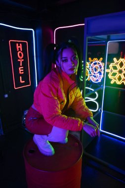 stylish young asian woman sitting near neon sign with hotel lettering outside 