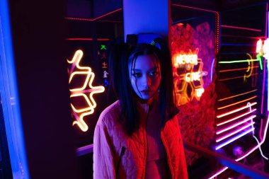 stylish young asian woman looking at camera near neon lighting outside 