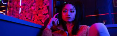young asian woman looking away near neon lighting, banner