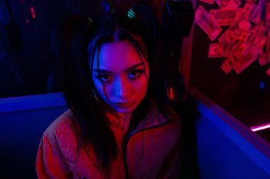 stylish young asian woman looking at camera near neon lighting 
