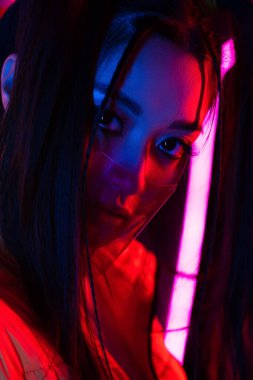young asian woman in face shield looking at camera near neon lighting 