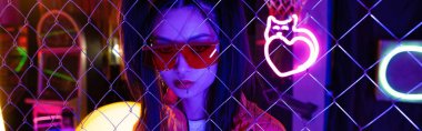 trendy young asian woman in red sunglasses near neon sign and metallic fence, banner