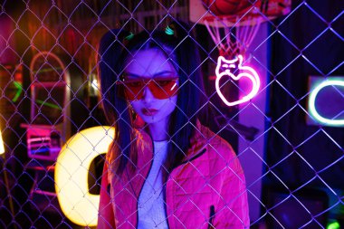 trendy young asian woman in red sunglasses near neon sign and metallic fence 