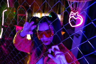 trendy asian woman in sunglasses near neon sign and metallic fence 