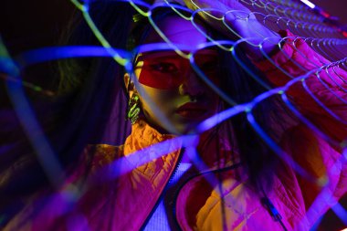 young asian woman in sunglasses looking at camera through metallic fence 