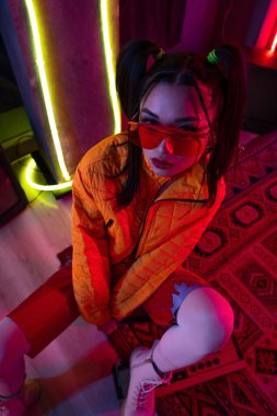 high angle view of stylish young asian woman in sunglasses and orange jacket posing near neon lighting 