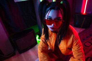 high angle view of stylish young asian woman in sunglasses and orange jacket posing near neon lighting 